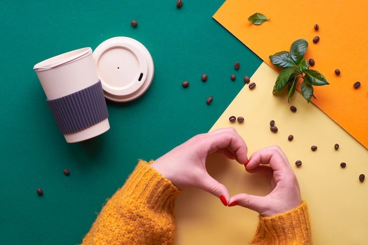 zero-waste-coffee-eco-friendly-reusable-coffee-cups-hands-in-orange-sweater-showing-heart-sign-geometric-flat-lay-on-split-tone-paper-geometric-with-beans-and-leaves_87646-3304.jpg